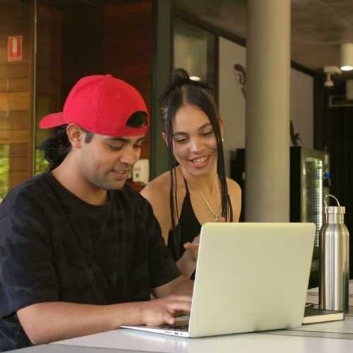 a young man and a young woman looking at a laptop, both smiling. The woman is supporting the young man. Performance coaching